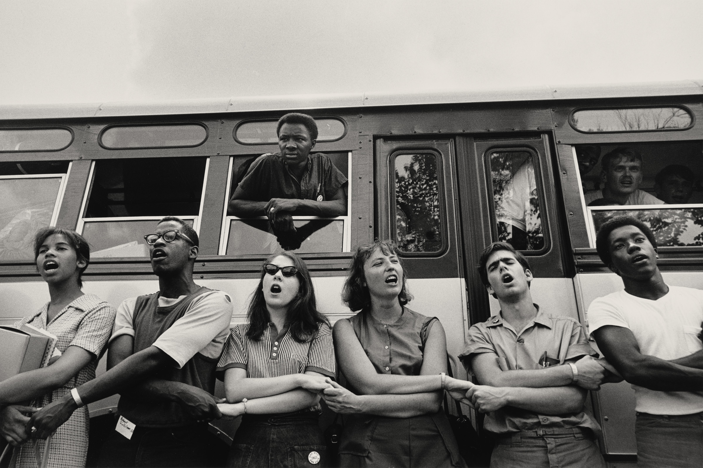 We Shall Overcome; Summer Freedom Bus, 1964. Photograph by Steve Schapiro. Freedom Summer volunteers form a chain with their arms while singing 'We Shall Overcome' before they get on the bus to Mississippi. Photograph in the collection of Miami University Art Museum, Oxford Ohio. Partial gift of the artist and partial purchase with contributions from the Kezur Endowment Fund (2019.23.1).