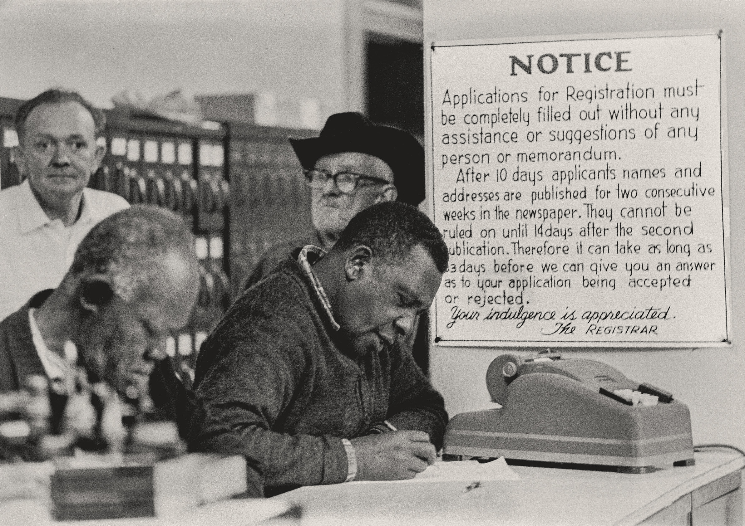 Threat to Registration, Hattiesburg, Mississippi, 22 January 1964. Photograph by Matt Herron. Black citizens filling out voter registration forms at the Forrest County Courthouse Clerk's Office run by Theron Lynd. Sign on the wall indicates the ordeal of public exposure applicants face, a tactic used to discourage Black registration. © 1976, Matt Herron / Photograph provided by TakeStock.